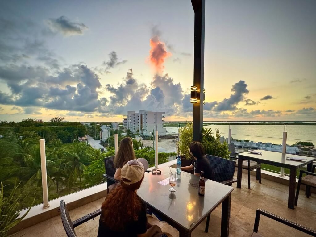 A terrace setting with people looking out over a sunset landscape with clouds and water.