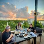A group enjoys an outdoor meal on a terrace overlooking lush greenery during a vibrant sunset.