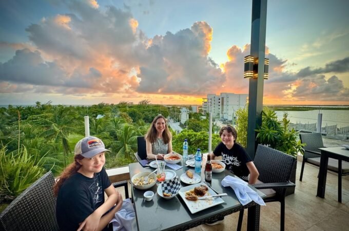 A group enjoys an outdoor meal on a terrace overlooking lush greenery during a vibrant sunset.