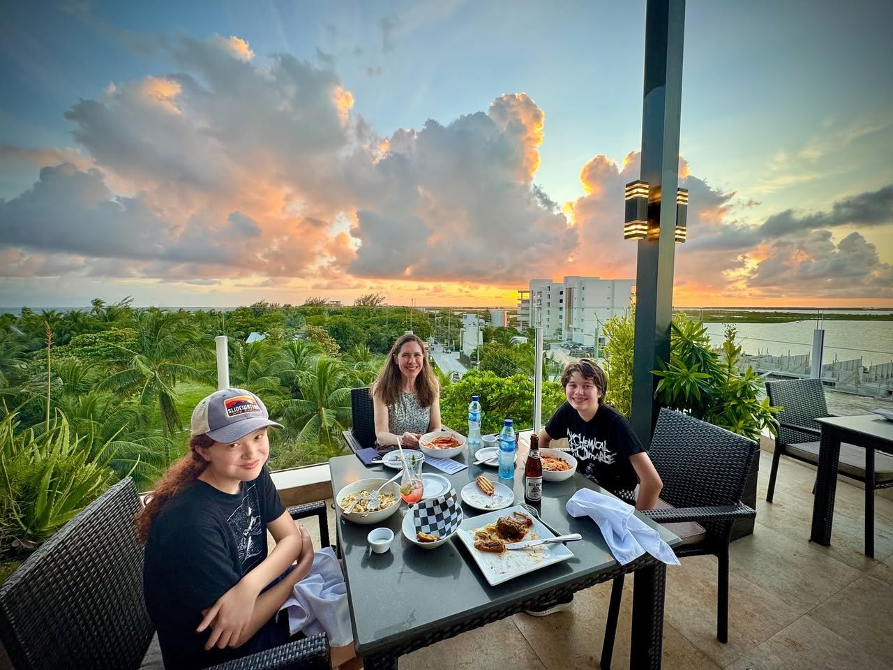 A group enjoys an outdoor meal on a terrace overlooking lush greenery during a vibrant sunset.
