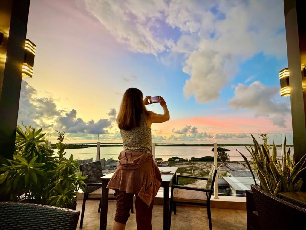 A person stands on a tiled terrace in Bali, photographing a colorful sunset sky filled with large cumulus clouds and ocean views.