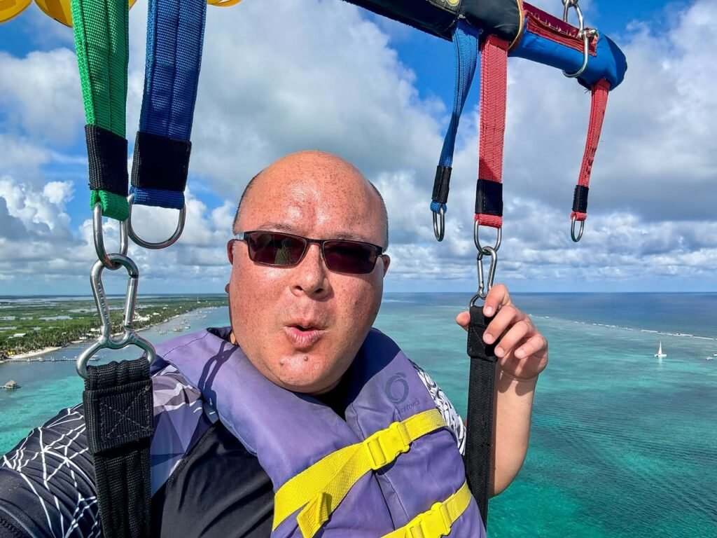 A passenger on a paraglider aloft over turquoise water with green and blue coastlines below.