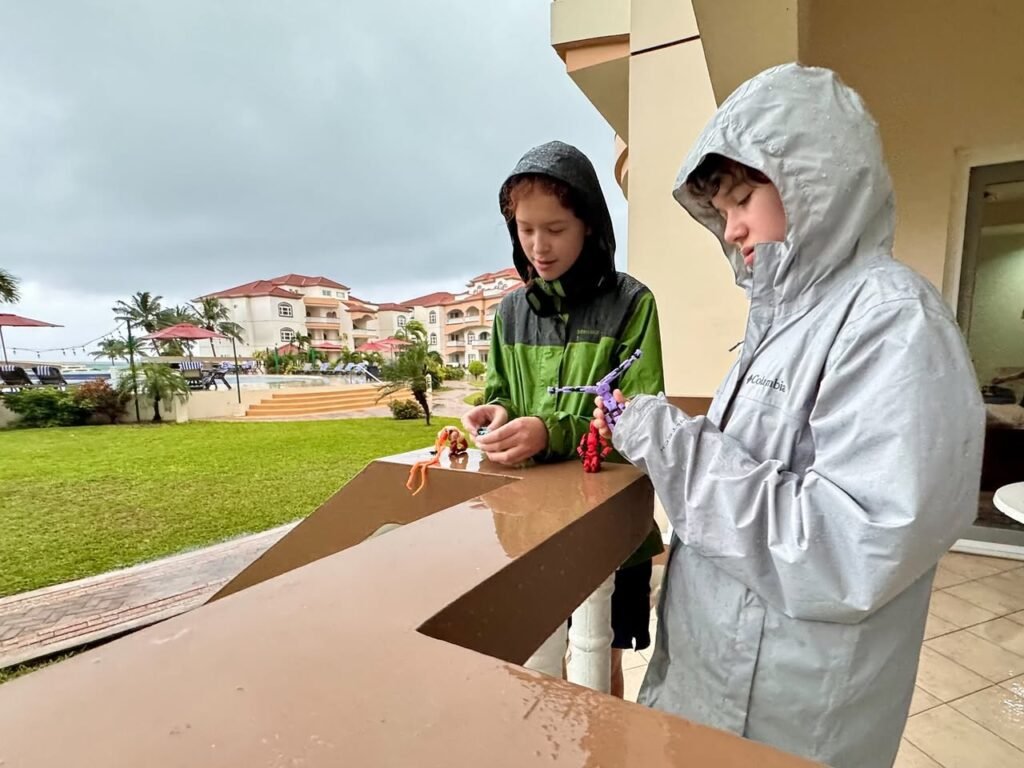 Two children wearing hoods and raincoats stand at a wet balcony railing during a rainstorm, with a view of a resort campus and manicured lawn under a cloudy sky.