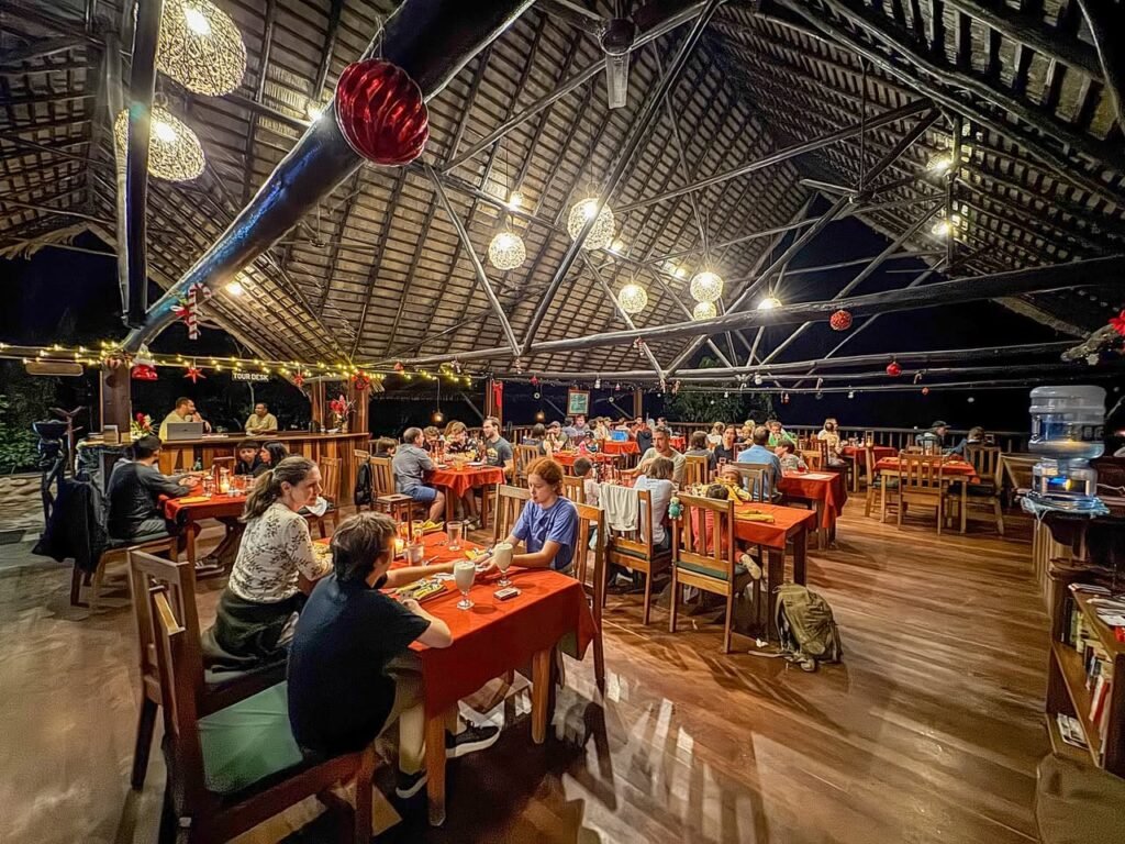 Diners at an outdoor restaurant under a wide banana leaf roof. The thatched ceiling is illuminated by hanging globe lights at dusk.