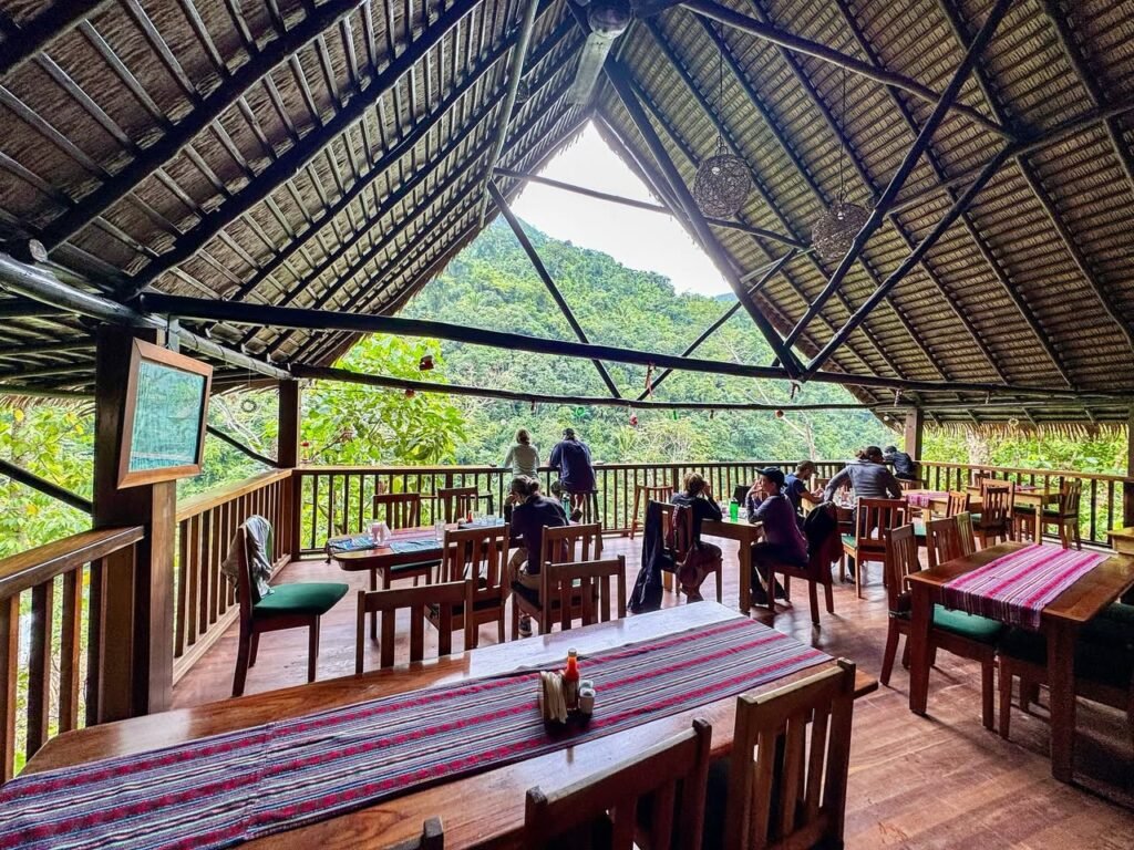 An open-air restaurant with wooden tables set under a bamboo roof. The structure has a large triangular opening, offering a scenic view of a lush green mountain below.