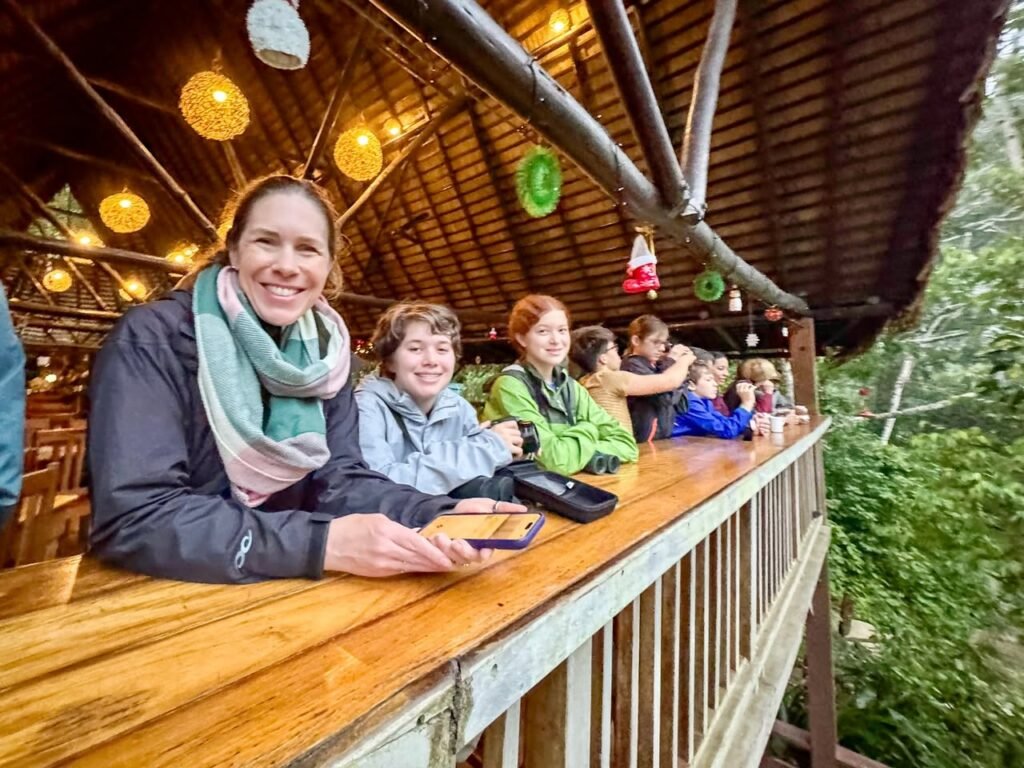 A group of people enjoys time on an elevated wooden walkway with a thatched roof and colorful lights.