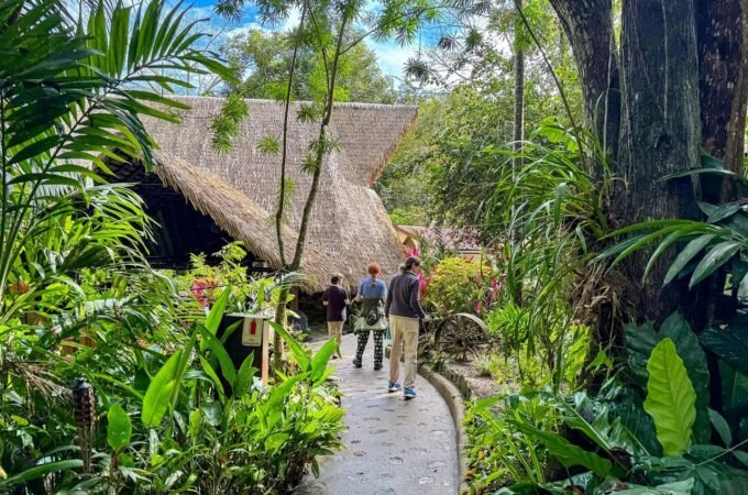 A stone path winds through a lush, green tropical garden toward a traditional thatched-roof building. Visitors stroll along the walkway amidst dense foliage and palm trees.
