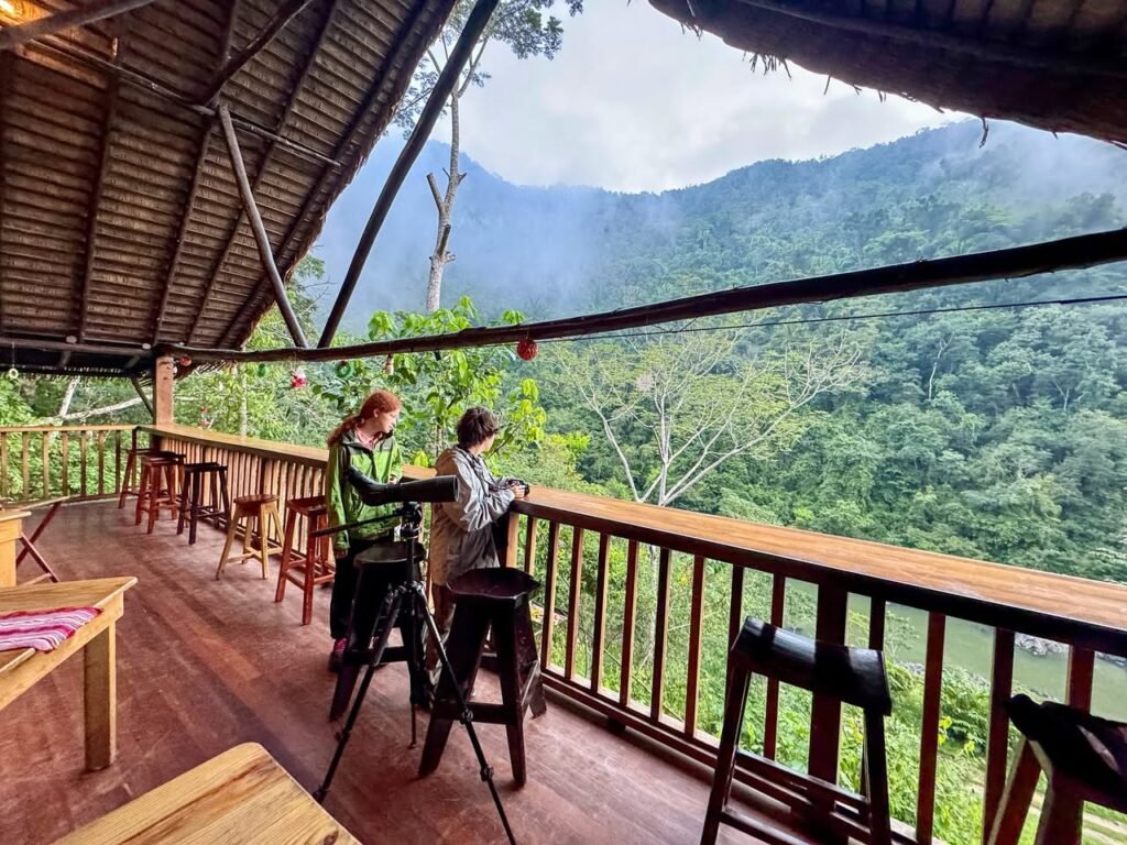 People standing on a wooden balcony overlooking lush green mountains under a striped tarp roof.