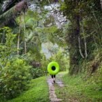 A person walks away down a green jungle path holding a large neon green ring.