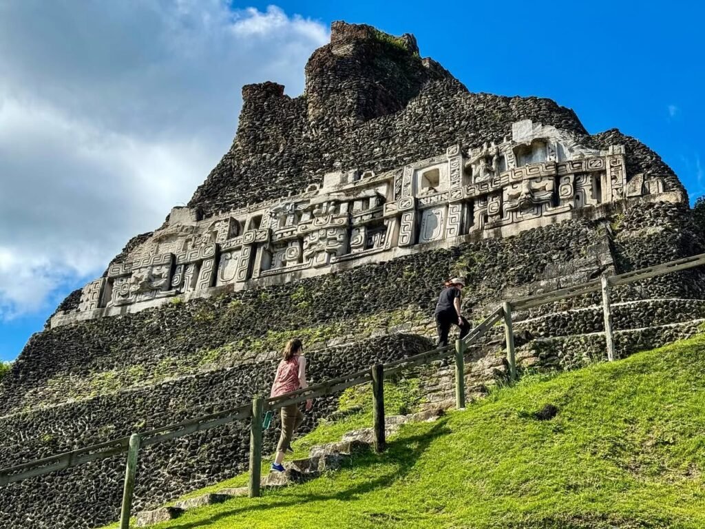A panoramic view of the Temple of the Sun at Machu Picchu, featuring stone ruins on a grassy hillside. Visitors walk along steps under a bright blue sky with scattered clouds.