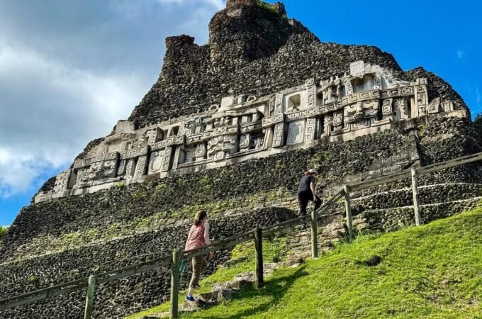 A panoramic view of the Temple of the Sun at Machu Picchu, featuring stone ruins on a grassy hillside. Visitors walk along steps under a bright blue sky with scattered clouds.