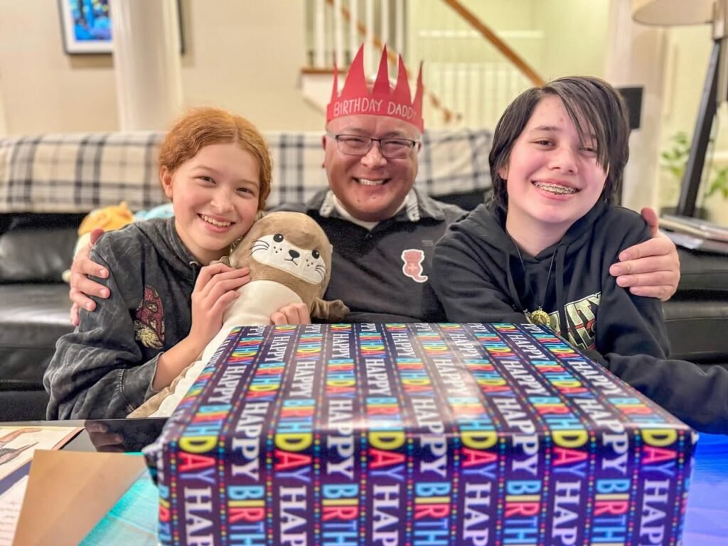 A happy family gathered around a colorful birthday cake with a stuffed animal in a home living room. The banner on the cake reads 'Happy Birthday'.