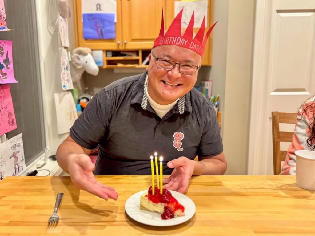 A person wearing a red spiky birthday paper crown sits at a table with a piece of chocolate cake topped with strawberries and lit candles on a white plate.