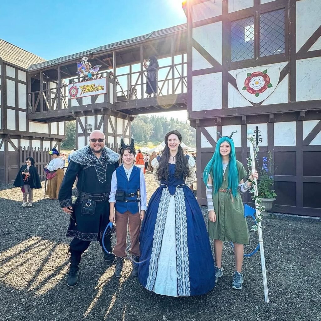 A family dressed in elaborate fantasy-themed costumes poses outside a Tudor-style building at a theme park. They stand on a gravel path with a whimsical backdrop.