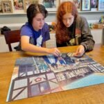 Two individuals collaborate on a large jigsaw puzzle depicting a traditional European street scene on a wooden dining table.