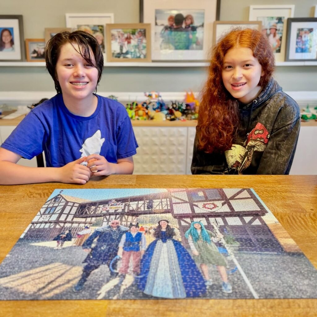 Two individuals sitting at a table with a mosaic artwork depicting a historical scene, presented against a backdrop of framed family photos and shelves.