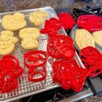 Renaissance Puzzle A close-up of freshly baked sugar cookies cooling on a wire rack with decorative red cutting shapes nearby.