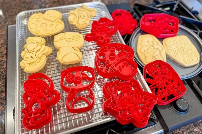 A close-up of freshly baked sugar cookies cooling on a wire rack with decorative red cutting shapes nearby.