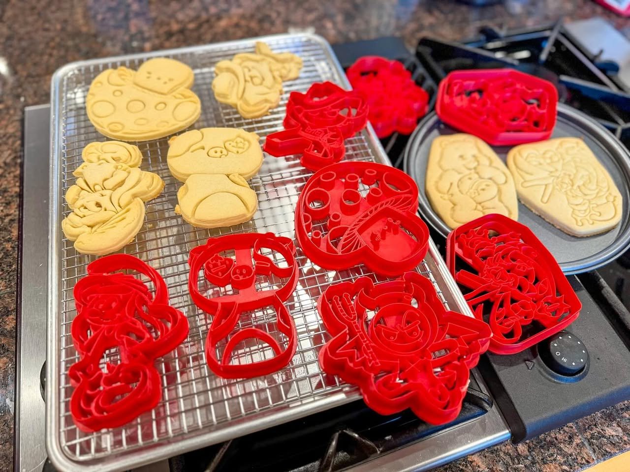 3D-Printed Cookie Cutters A close-up of freshly baked sugar cookies cooling on a wire rack with decorative red cutting shapes nearby.