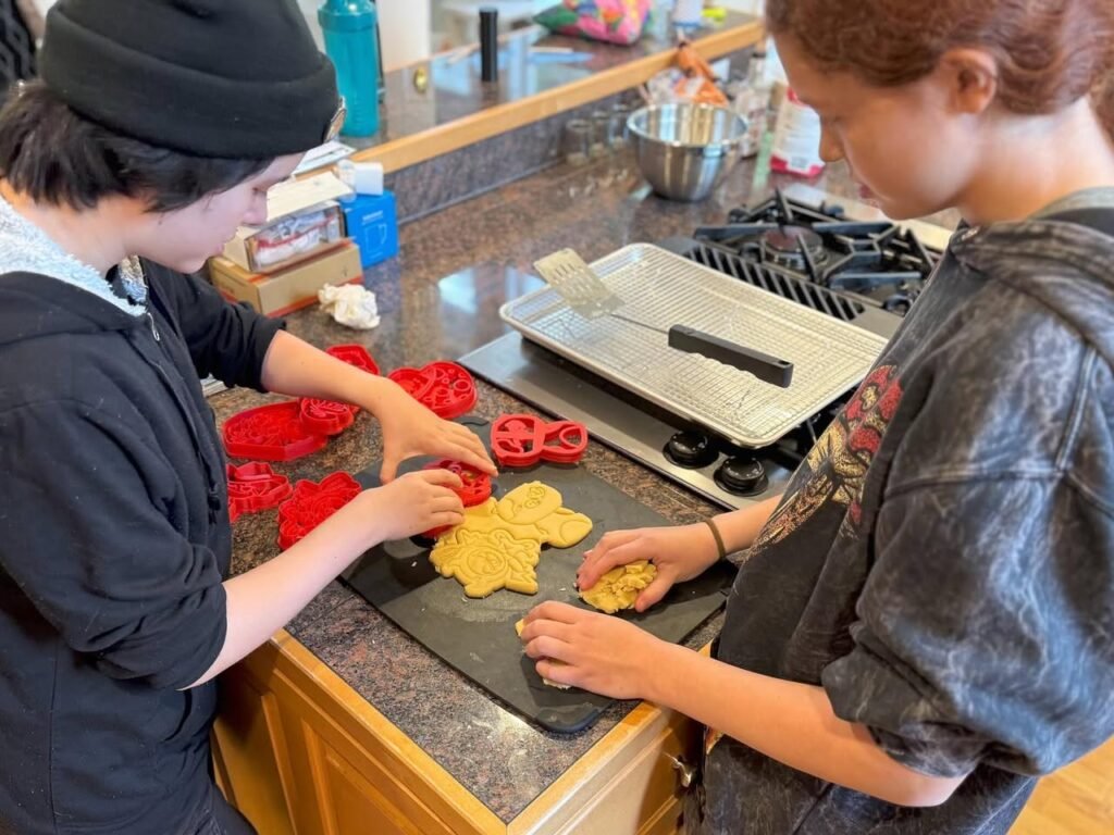 Two people in a home kitchen using red cookie cutters to shape yellow dough into leaf shapes on a dark board, with a metal baking tray on the stove.