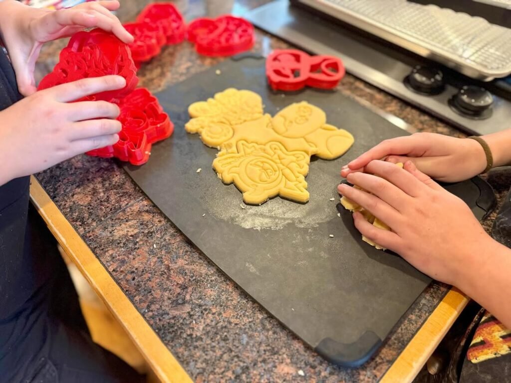 Hands using red cookie cutters on rolled dough on a dark cutting board to make holiday cookies.