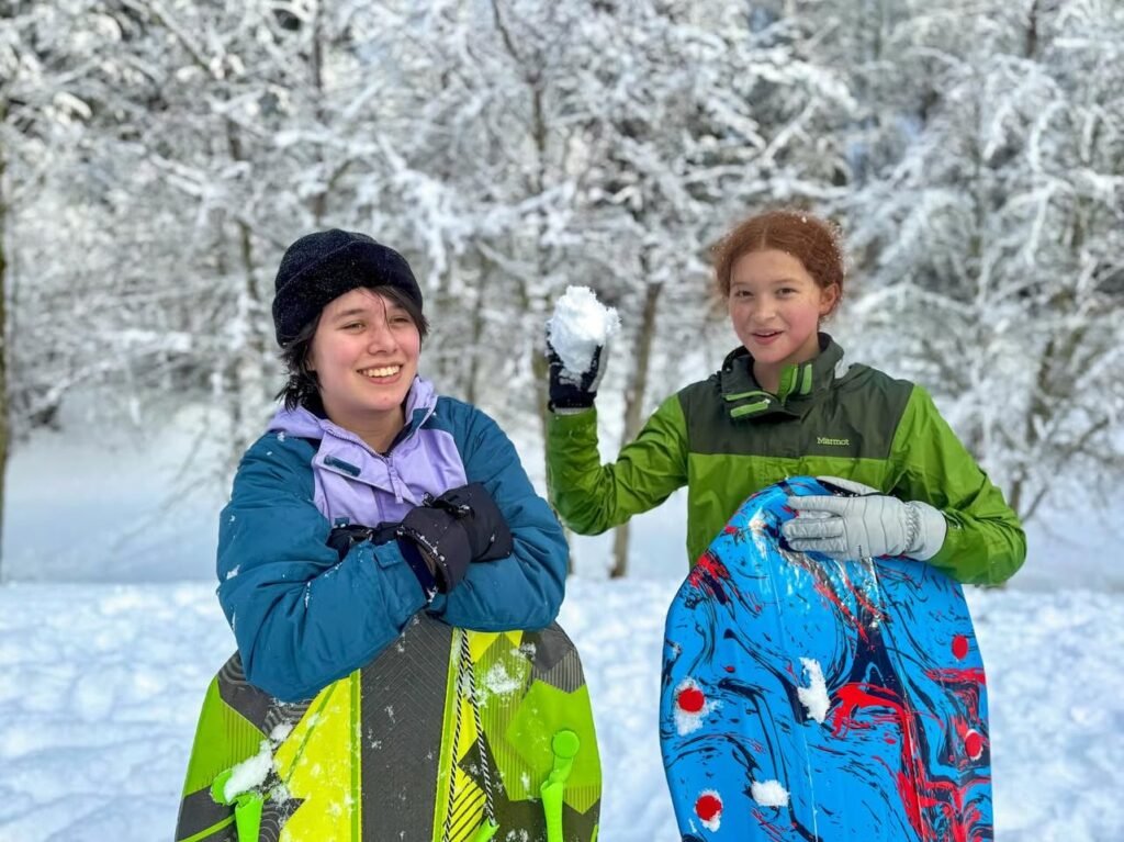 Two individuals pose with snowboards in a winter forest, one holding a snowball. The background features trees heavily laden with snow.