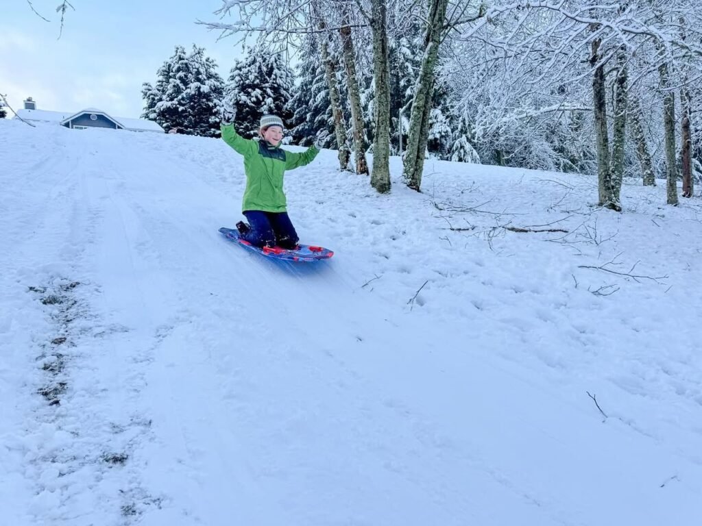 A child sleds down a snowy hill with arms raised, surrounded by frosted trees in the Pacific Northwest