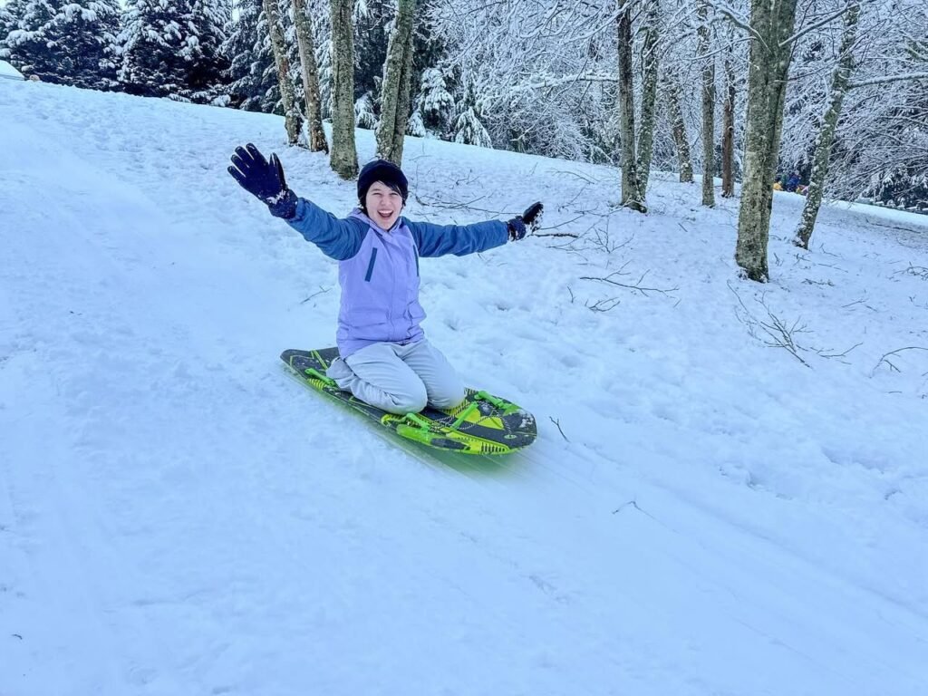 A person sledding down a snowy hill in the Pacific Northwest. Snow-covered trees surround the winter landscape.
