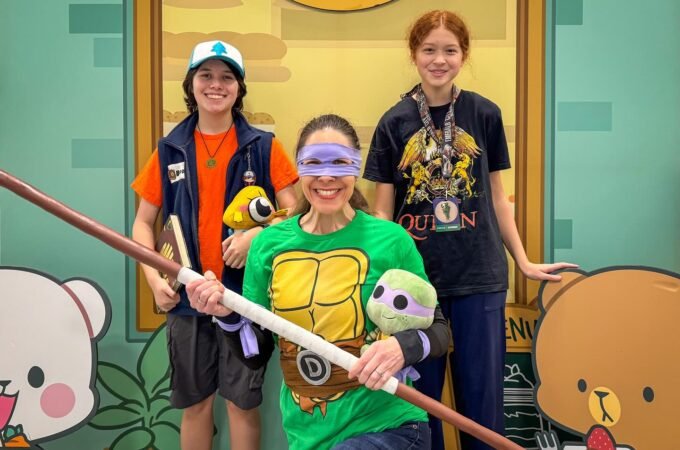 Three people in playful costumes and accessories posing with a prop stick against a bright cartoon-style background featuring a bear and a pig.