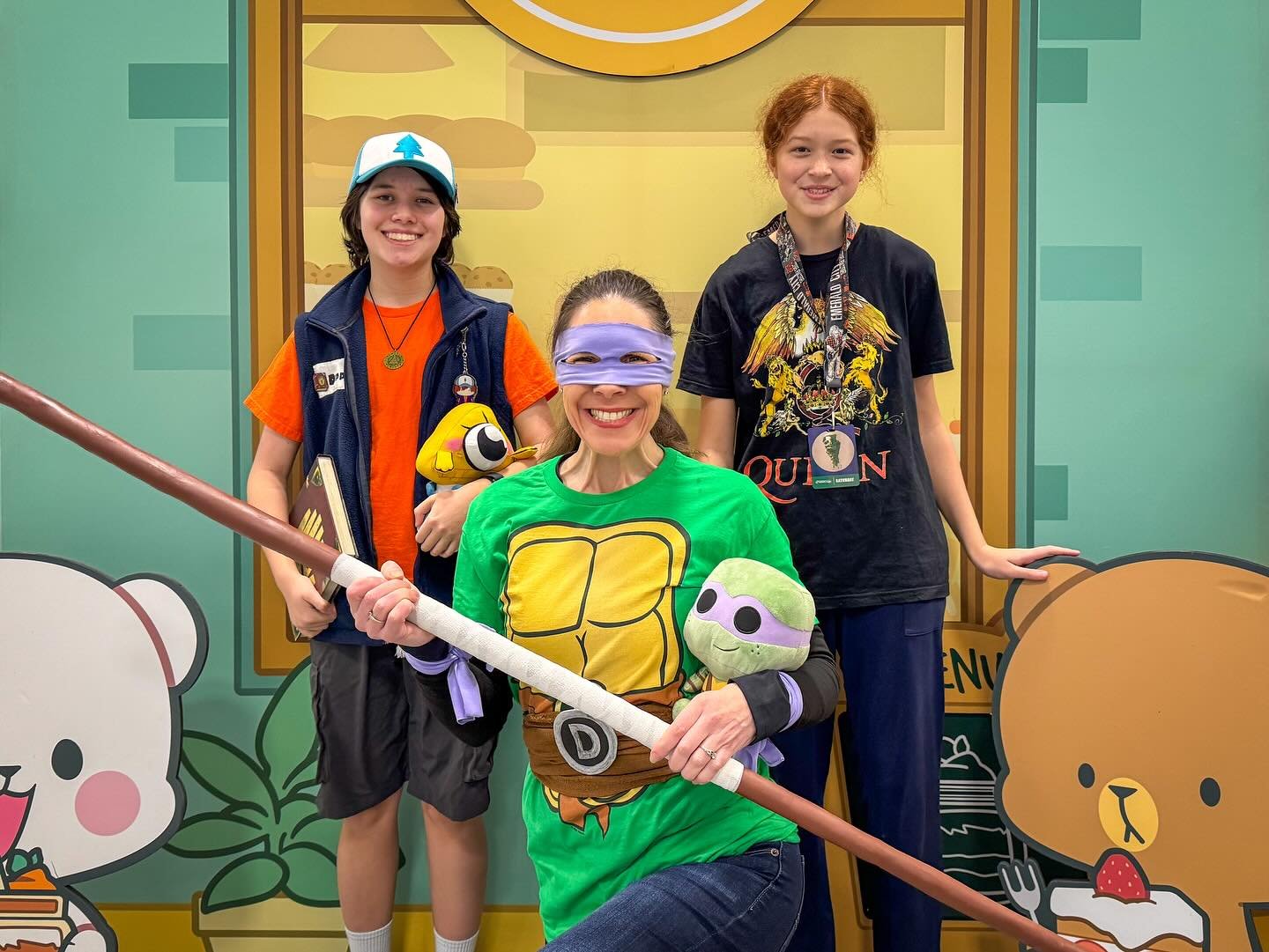 Three people in playful costumes and accessories posing with a prop stick against a bright cartoon-style background featuring a bear and a pig.
