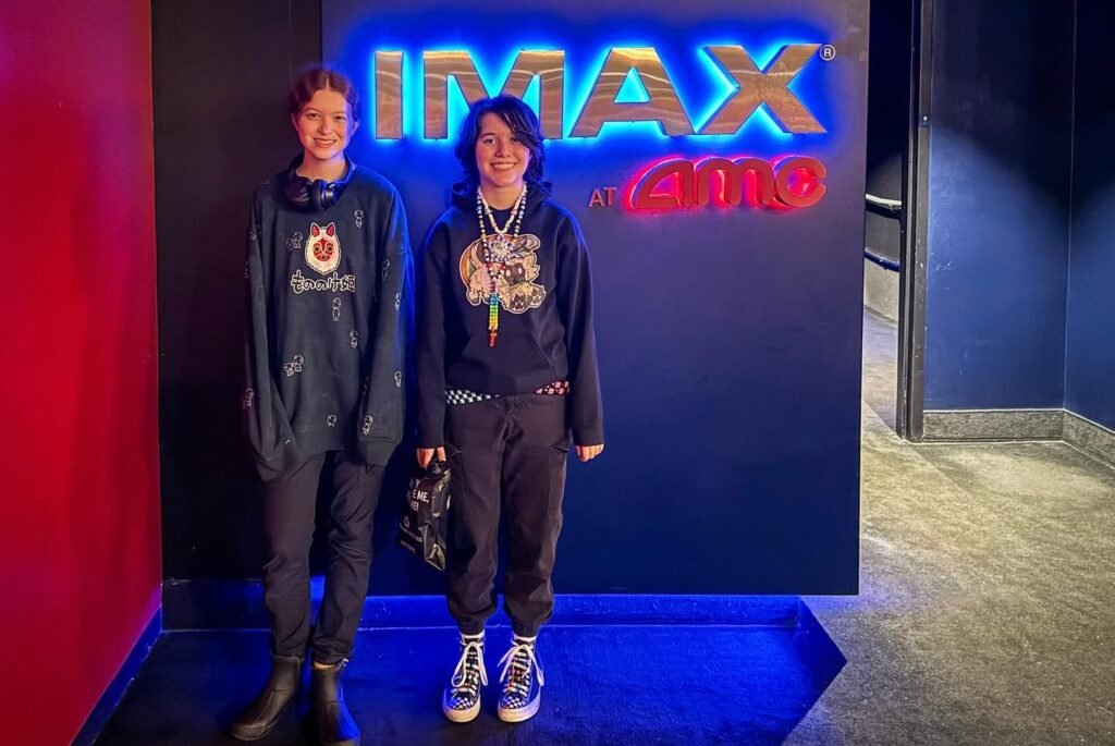 Two friends posing in front of a bright IMAX logo at a modern movie theater.