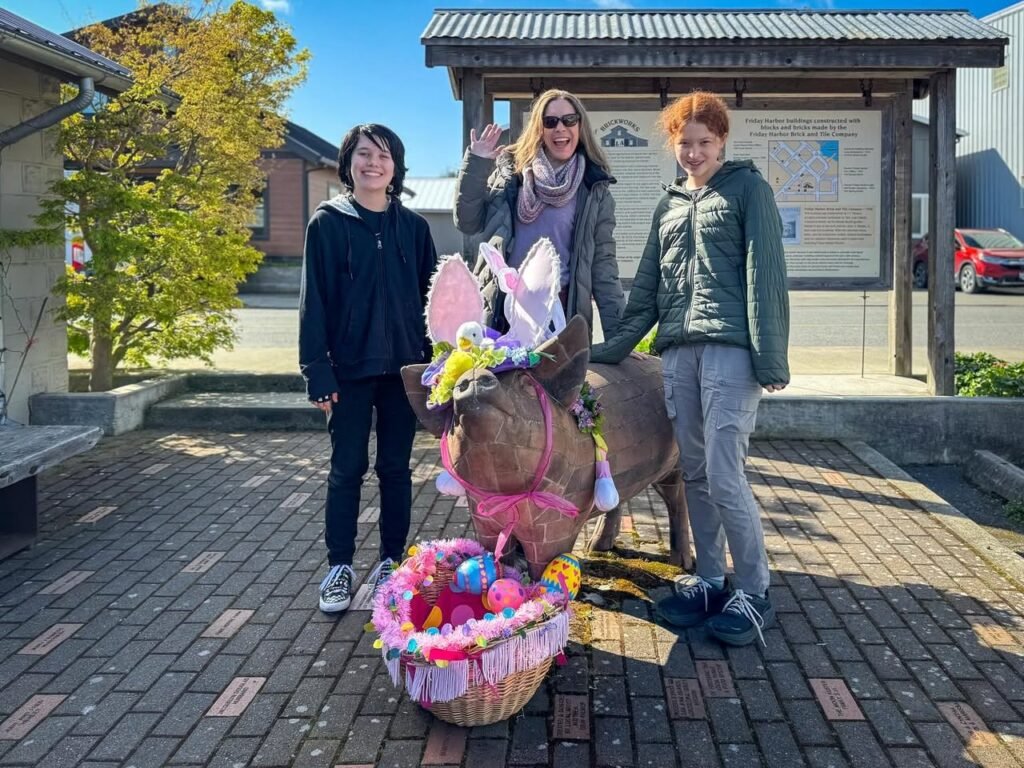 Three people standing beside a donkey decorated with Easter eggs and a festive basket. One person waves from a wooden shelter on a bright, sunny day.