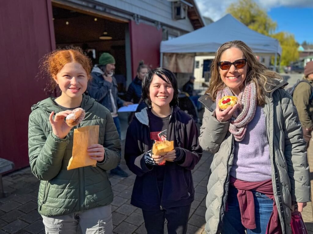 Three people holding pastries outside a red building on a sunny day. The background shows a market tent and autumn trees.