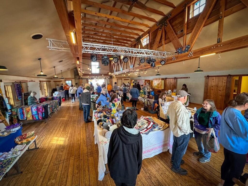 Visitors browse tables at an indoor community craft fair. The scene features a high wooden ceiling with trusses in a warm, rustic hall.