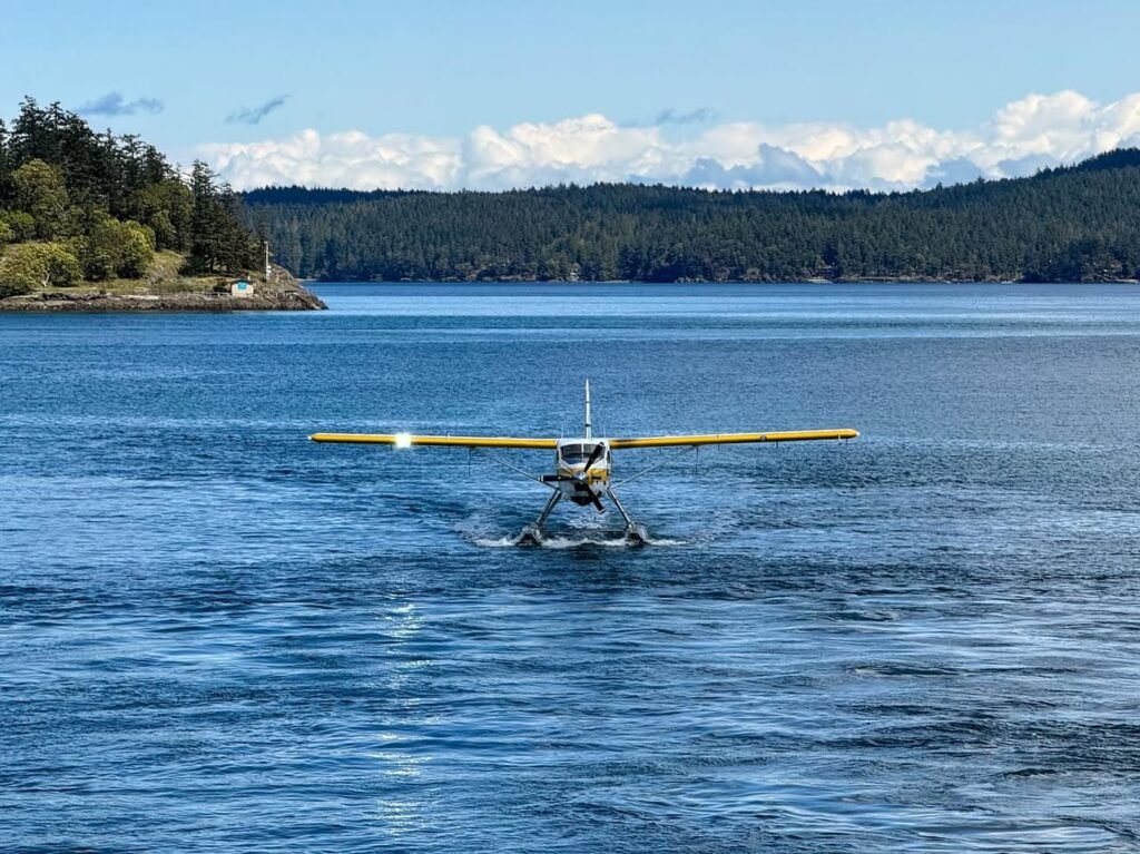 A yellow seaplane taxis across calm blue waters with evergreen islands in the background.