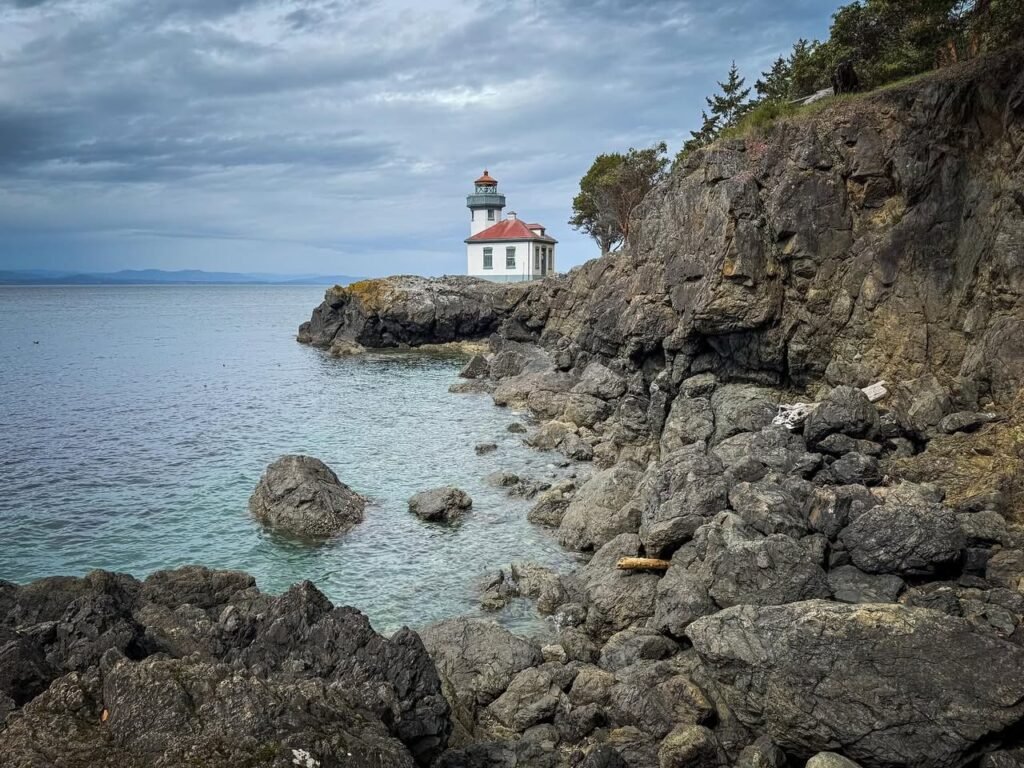 A red-roofed lighthouse stands on a rocky headland beside calm turquoise water under a cloudy sky.
