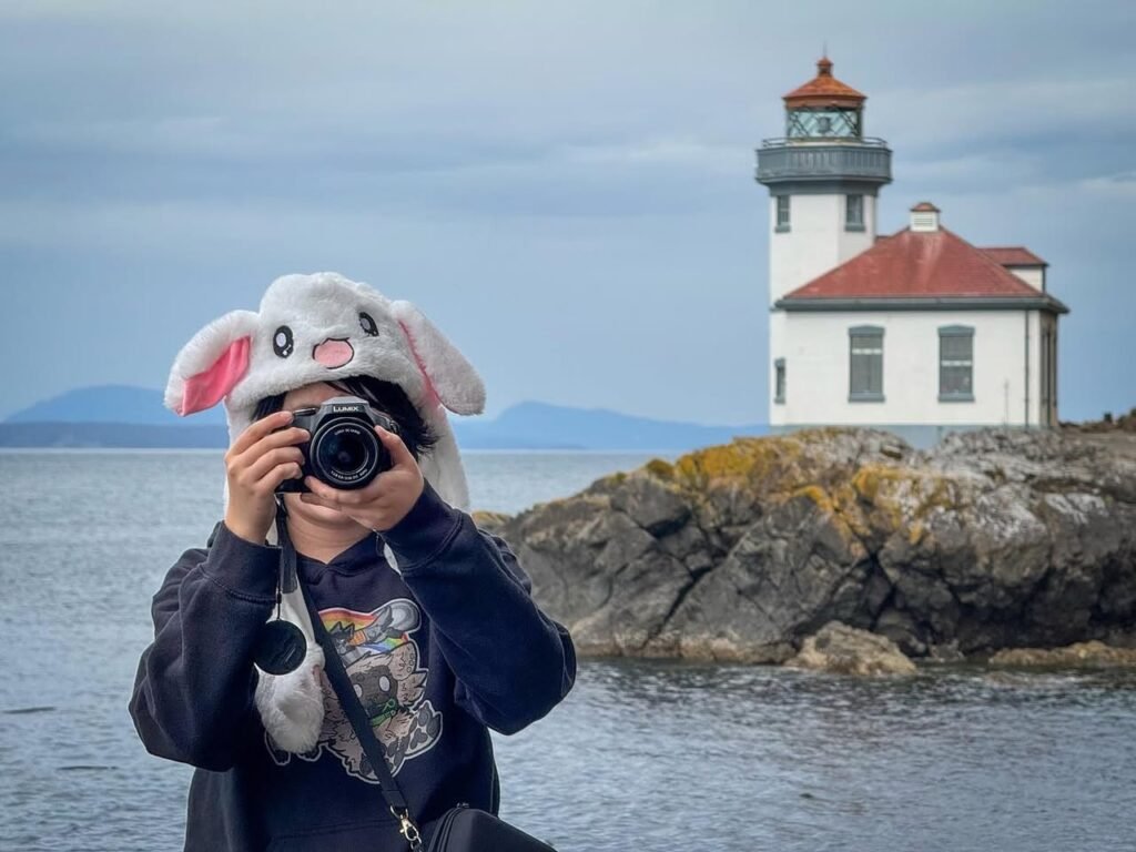 A person wearing a white bunny hat taking a photo near a white lighthouse on rocky coast. The lighthouse has a red roof and is surrounded by water and hills.