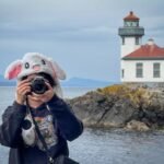 A person wearing a white bunny hat taking a photo near a white lighthouse on rocky coast. The lighthouse has a red roof and is surrounded by water and hills.