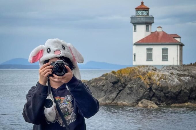 A person wearing a white bunny hat taking a photo near a white lighthouse on rocky coast. The lighthouse has a red roof and is surrounded by water and hills.