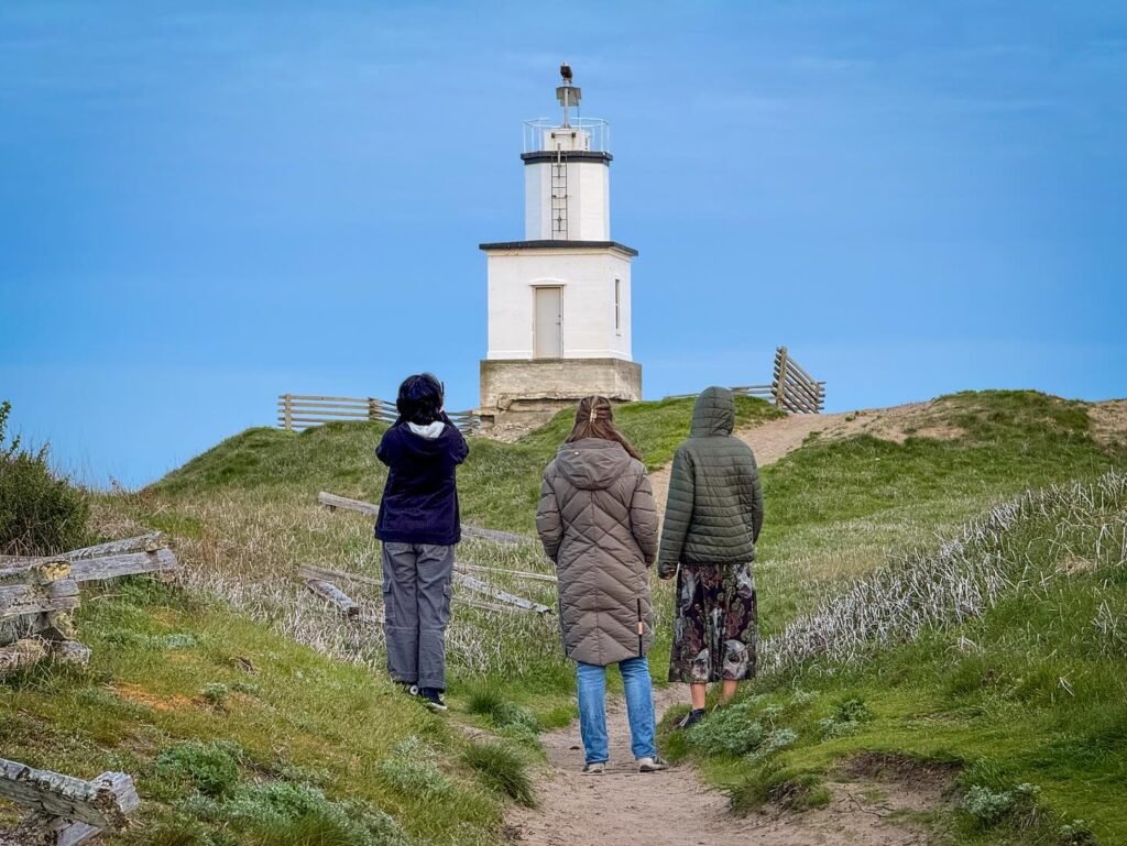 Three people walk up a grassy hill toward a white lighthouse under a clear blue sky.