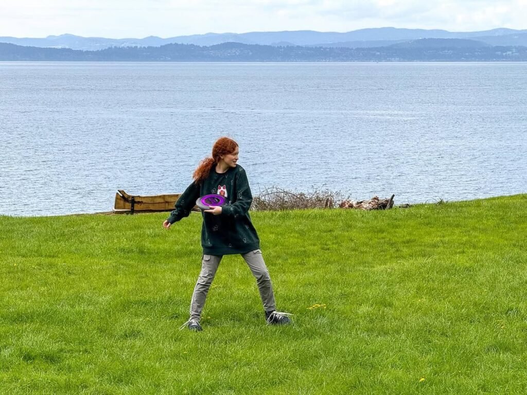 A person stands on a grassy slope holding a purple frisbee, with a large body of water and distant mountains in the background.