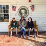 A family sits on a wooden bench in front of the West Virginia State Capitol building with state flags and a seal displayed on the wall.