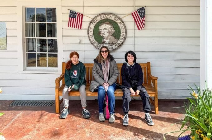 A family sits on a wooden bench in front of the West Virginia State Capitol building with state flags and a seal displayed on the wall.