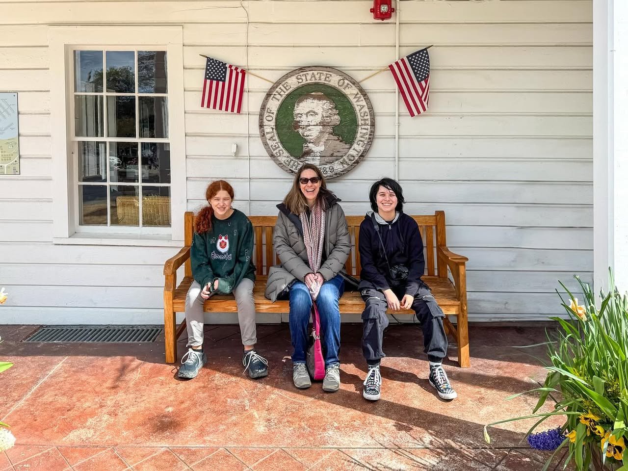 A family sits on a wooden bench in front of the West Virginia State Capitol building with state flags and a seal displayed on the wall.