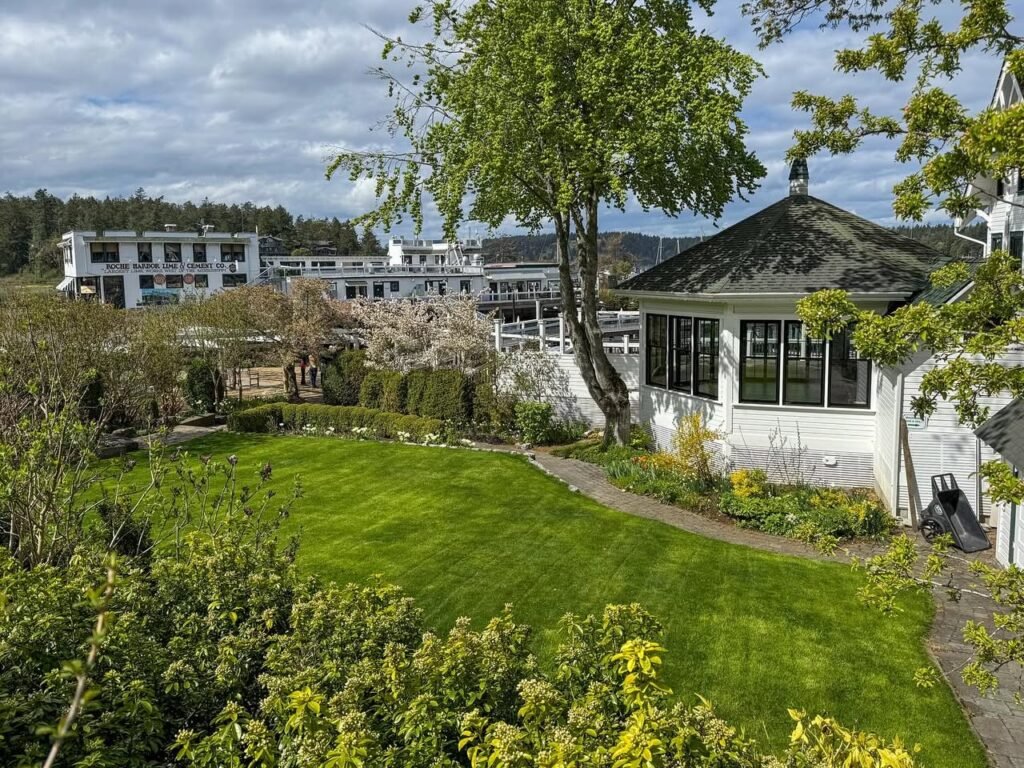 A scenic garden view featuring lush green lawns, blooming trees, and a white building in the background under a cloudy sky.