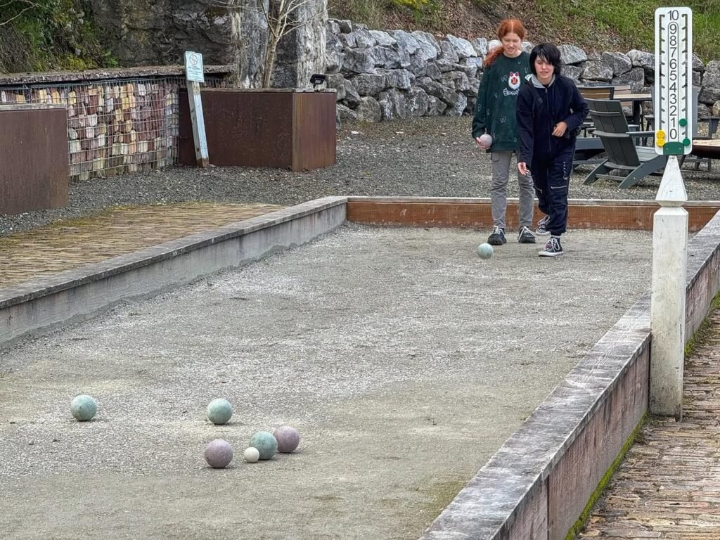 Two people standing at the end of a gravel bocce court as a ball rolls toward the target.