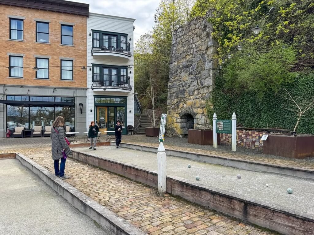 A city plaza features modern townhomes and a historic stone stone ruins. Visitors walk along paved pathways near distinctive brick buildings.