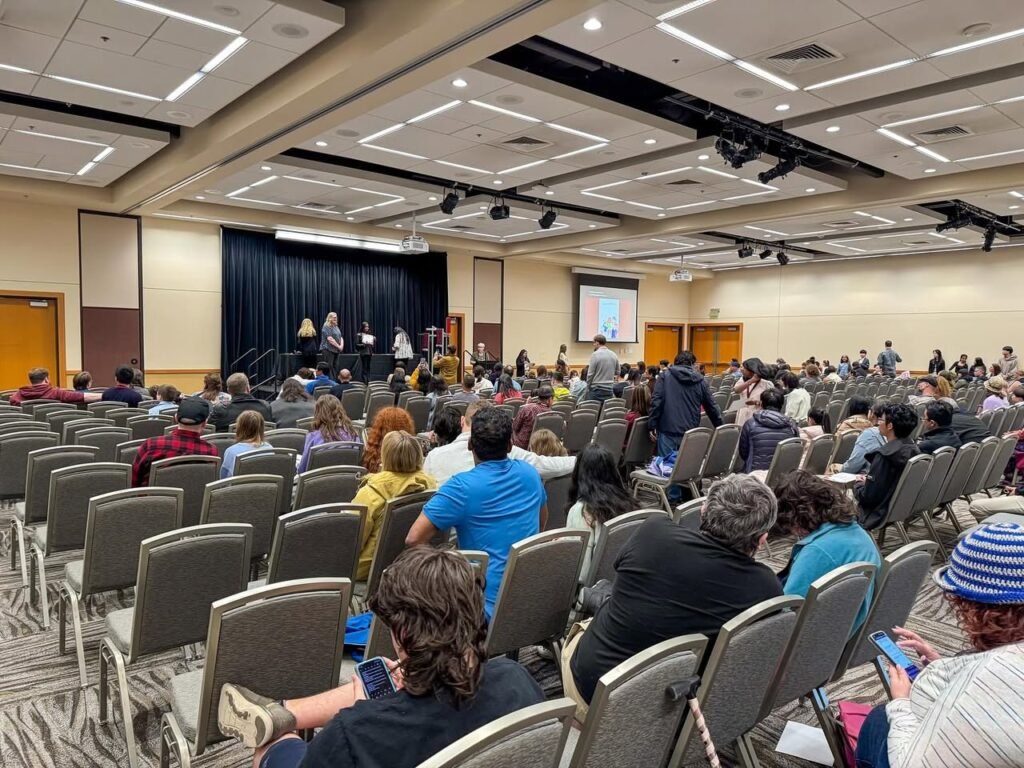 Attendees seated in rows facing a presentation screen in a large auditorium. A professional setting for a workshop or meeting.