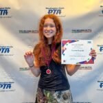 A young person with long red hair smiling, wearing a medal, and holding a certificate in front of a Washington State PTA backdrop.