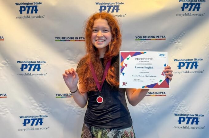 A young person with long red hair smiling, wearing a medal, and holding a certificate in front of a Washington State PTA backdrop.